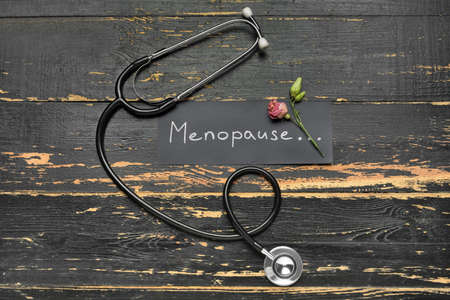 Paper with word MENOPAUSE, dried rose and stethoscope on dark wooden background