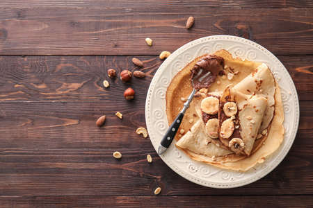 Plate of tasty thin pancakes with chocolate paste, cut banana and nuts on wooden backgroundの写真素材
