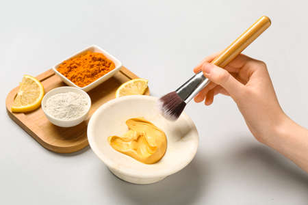Woman with makeup brush taking turmeric mask from bowl on gray backgroundの写真素材