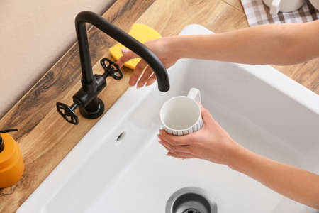 Woman pouring water from faucet onto cup near white sink