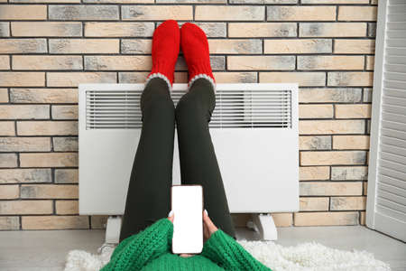 Woman in knitted socks with mobile phone warming feet at radiator. Concept of heating seasonの写真素材