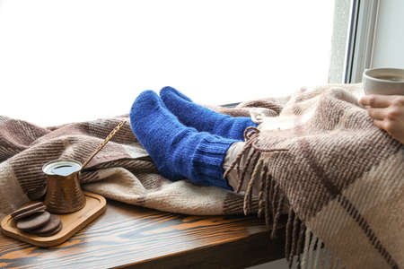 Woman in warm socks with cup of coffee holding legs on windowsill. Concept of heating seasonの写真素材