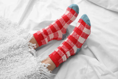 Female legs in knitted socks covered with plaid on bed, closeup. Concept of heating season