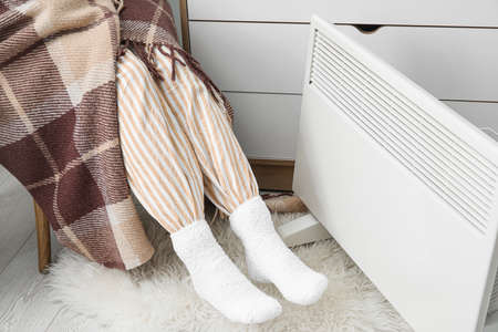 Woman in soft socks sitting in chair and warming feet near radiator. Concept of heating season