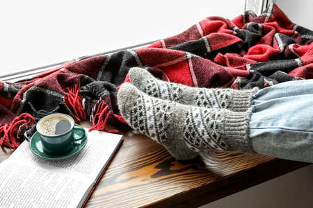 Female legs in warm socks, cup of coffee and opened book on windowsill. Concept of heating seasonの写真素材