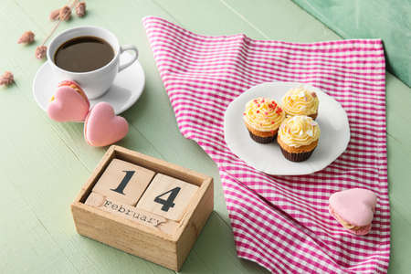 Calendar with date of Valentines Day, cup of coffee and tasty cupcakes on green background