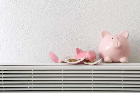 Whole and broken piggy bank with coins on radiator near white wall. Concept of heating seasonの写真素材