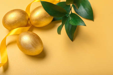 Golden Easter eggs, plant branch and ribbon on color background, closeup
