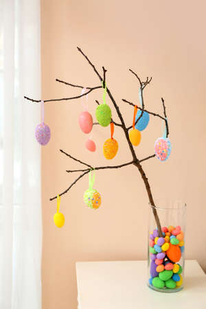 Vase with tree branch and different Easter eggs on table near light wall