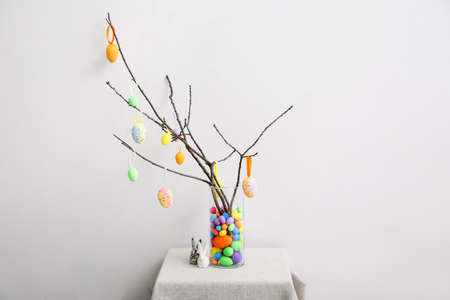 Tree branches with Easter eggs and small toy rabbits on table near light wall
