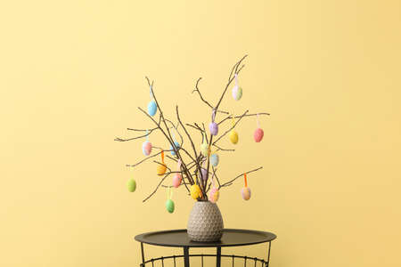 Vase with tree branches and Easter eggs on table against color wall