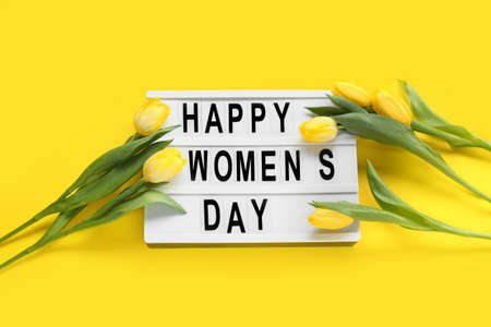 Board with text HAPPY WOMEN'S DAY and tulips on yellow background