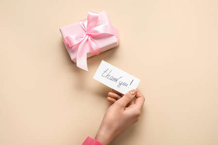 Female hand holding sheet of paper with text THANK YOU and gift box on beige backgroundの写真素材