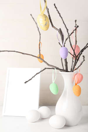 Vase with tree branches, Easter eggs and blank photo frame on white wooden background, closeup