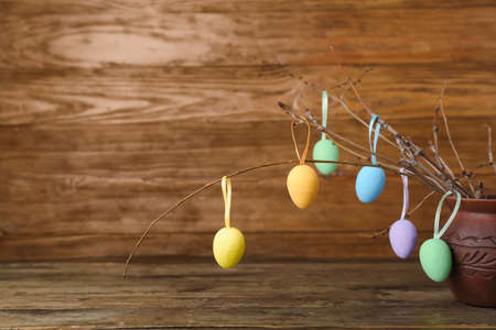 Clay vase with tree branches and different Easter eggs on wooden background, closeup
