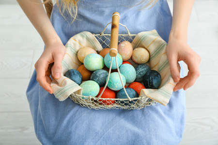 Woman holding basket with painted Easter eggs on light background