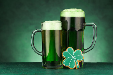 Glasses of beer and gingerbread cookies for St. Patrick's Day celebration on green background