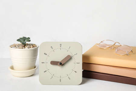Alarm clock, stack of books, eyeglasses and houseplant on table near light wall