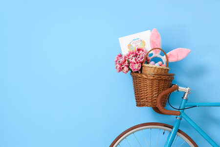 Bicycle with baskets, tulips, Easter eggs, bunny ears and greeting card near blue wall