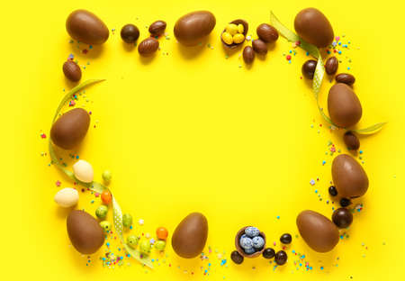 Frame made of chocolate Easter eggs, different candies and sprinkles on yellow background