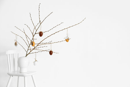 Vase with tree branches and Easter eggs on chair near light wall
