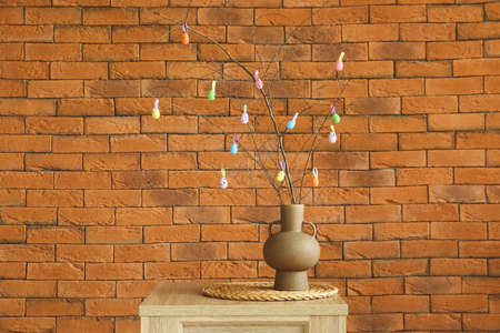 Vase with tree branches and colorful Easter eggs on table near brick wall