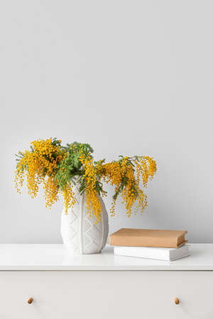 Vase with mimosa flowers and books on chest of drawers near light wallの写真素材