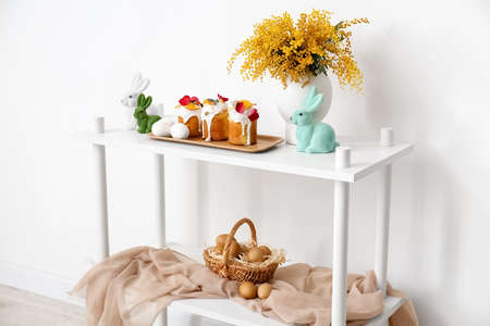 Tray with Easter cakes, eggs and mimosa flowers on shelf unit near white wall