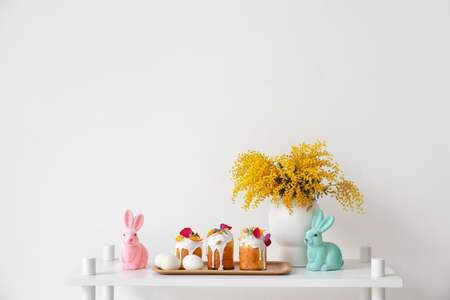 Tray with Easter cakes, eggs, decor and vase with mimosa flowers on shelf near white wallの写真素材