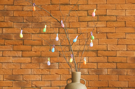 Vase with tree branches and colorful Easter eggs near brick wall
