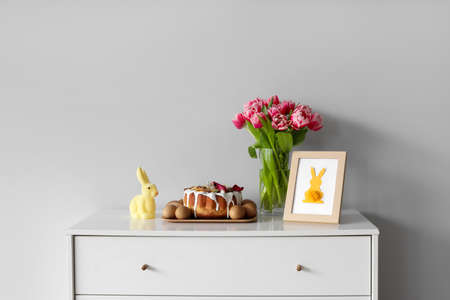 Chest of drawers with Easter cake, eggs, decor and flowers near light wall