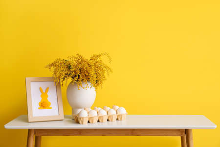 Vase with mimosa flowers, photo frame and holder with Easter eggs on table near color wall