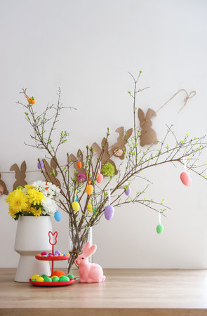 Stand with Easter eggs, beautiful flowers and stylish decor on counter near white wall