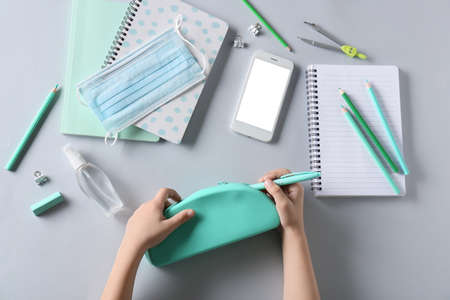 Child's hands with pencil case, medical mask, mobile phone and stationery on white background