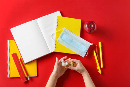 Child's hands with bottle of sanitizer, medical mask, stationery and apple on red background