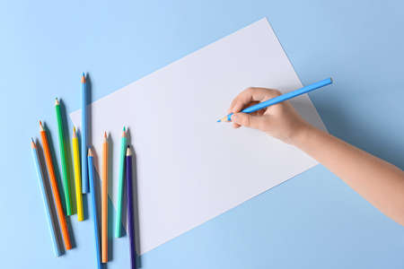 Child's hand with blank sheet of paper and different pencils on color background