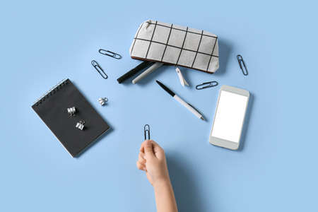 Child's hand with paper clip, mobile phone and stationery on color background