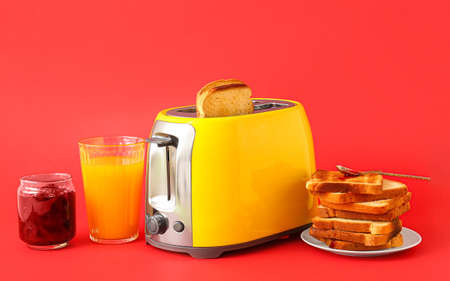 Modern toaster with bread slices, glass of juice and jam on red backgroundの写真素材