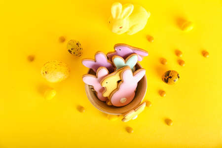 Bowl with tasty Easter cookies in shape on bunny, eggs and candies on yellow background