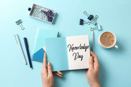 Female hands holding open notebook with written text KNOWLEDGE day and set of stationery on blue backgroundの写真素材