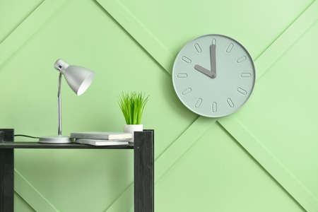 Table with books, houseplant, lamp and modern clock hanging on color wall in roomの写真素材
