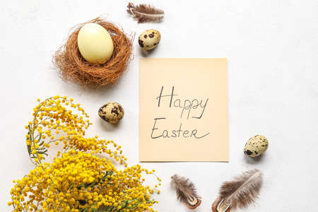 Easter composition with greeting card, eggs, feathers and mimosa branches on light background