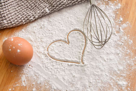 Fresh chicken egg, flour with drawn heart and whisk on wooden background