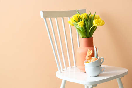 Vase with tulips and cups with Easter rabbits on chair near color wall