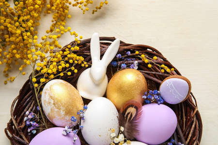Nest with painted Easter eggs, bunny and flowers on light wooden background, closeup