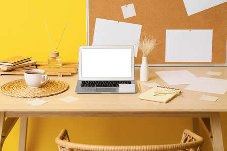Modern workplace with laptop, sticky notes, vase and cup of tea near yellow wall