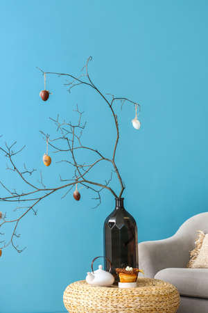 Vase with tree branches decorated for Easter celebration, delicious bun and teapot on table near blue wallの写真素材