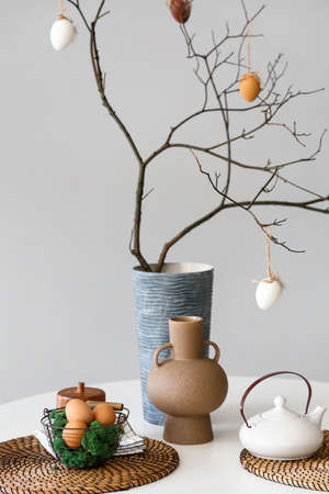 Basket with Easter eggs, teapot and vase with tree branches on dining table near light wallの写真素材