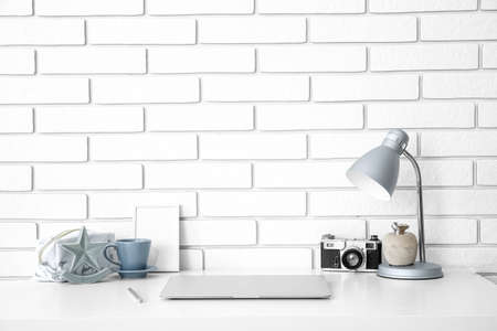 Workplace with laptop, lamp, photo camera and cup near white brick wall