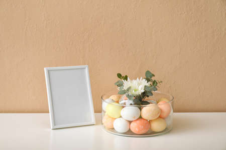 Bowl with Easter eggs, floral decor and blank photo frame on table near color wallの写真素材